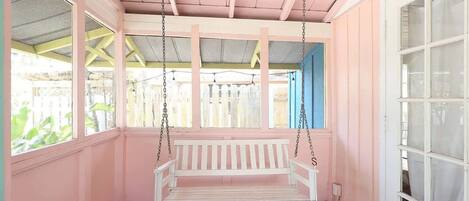A pink-walled porch with a white swing bench, a tiled floor, and a view of a wooden fence.