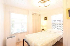 A bedroom with a bed, a nightstand with a lamp, a window with blinds, and a ceiling light fixture.