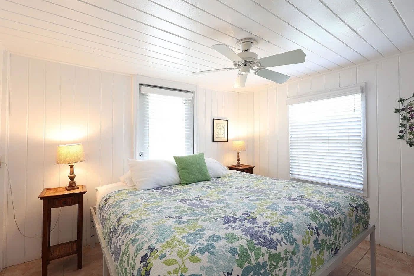 A bedroom with a bed, two bedside tables, a ceiling fan, and two windows with blinds.