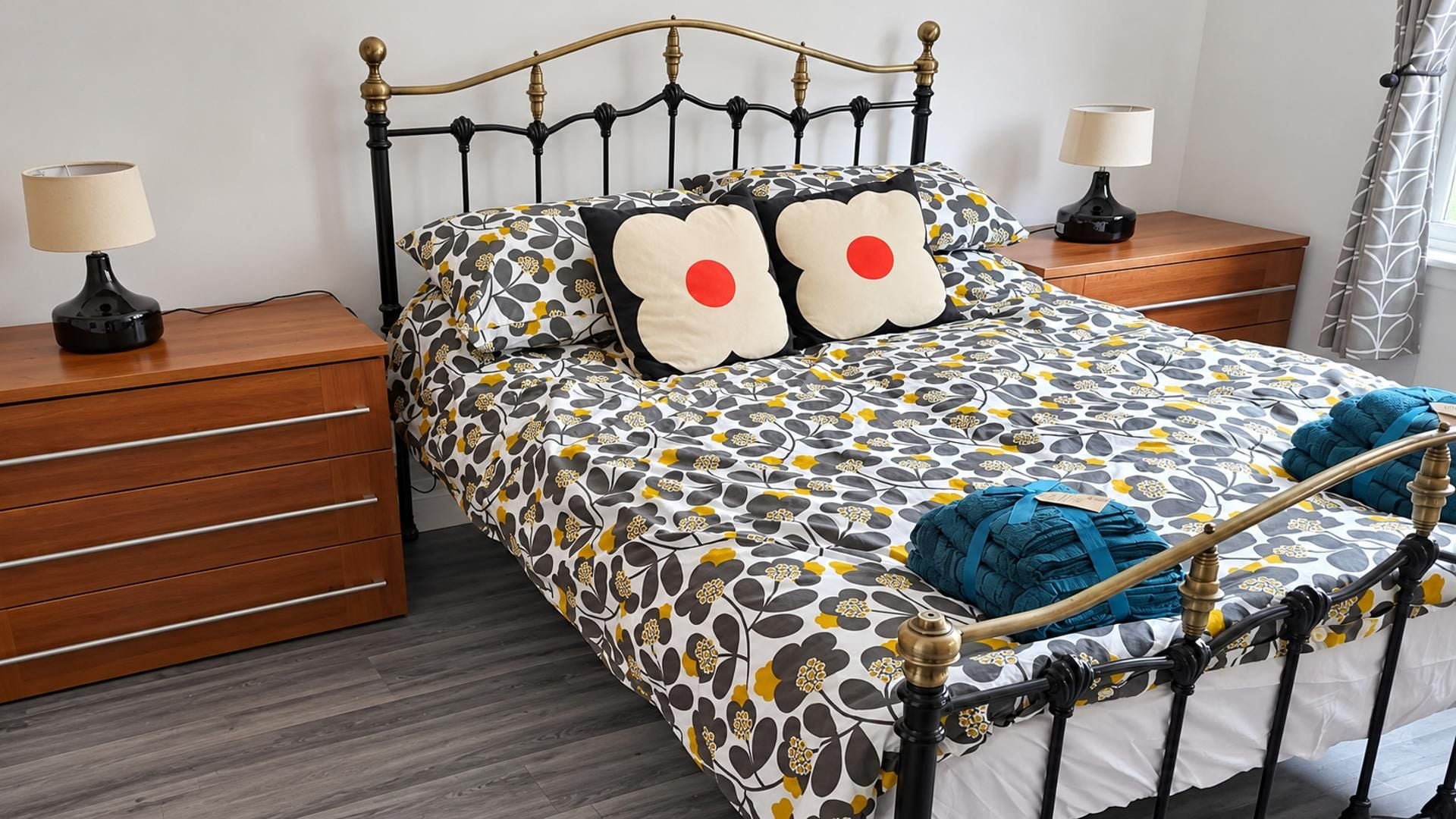 Uma cama com colcha estampada de flores, dois travesseiros com círculos vermelhos, um criado-mudo de madeira com uma luminária e um cobertor azul sobre a cama.