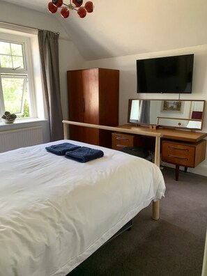 A hotel room with a bed, a desk with a chair, a wardrobe, a television, and a window with curtains. - Large home, 6 bedrooms, private parking, gardens (Barnstaple)