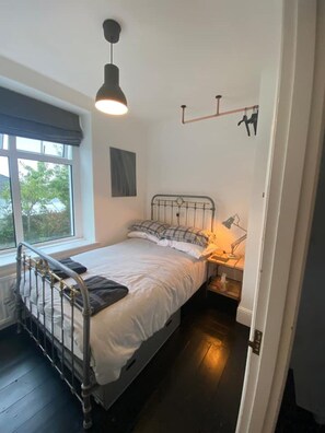 A small, single-bed room with a metal bed frame, a bedside table with a lamp, and a window with a view of trees. - Large home, 6 bedrooms, private parking, gardens (Barnstaple)