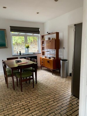 A dining area with a brick floor, a wooden table with chairs, a window with a view of greenery, and a cabinet with glassware. - Large home, 6 bedrooms, private parking, gardens (Barnstaple)