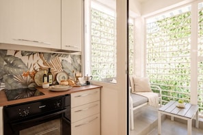 A modern kitchen with a built-in oven, a dining area with a bench, and a view through a window with a green trellis. - Ravanas City Escape | Stylish 2BR Apartment (Bari)