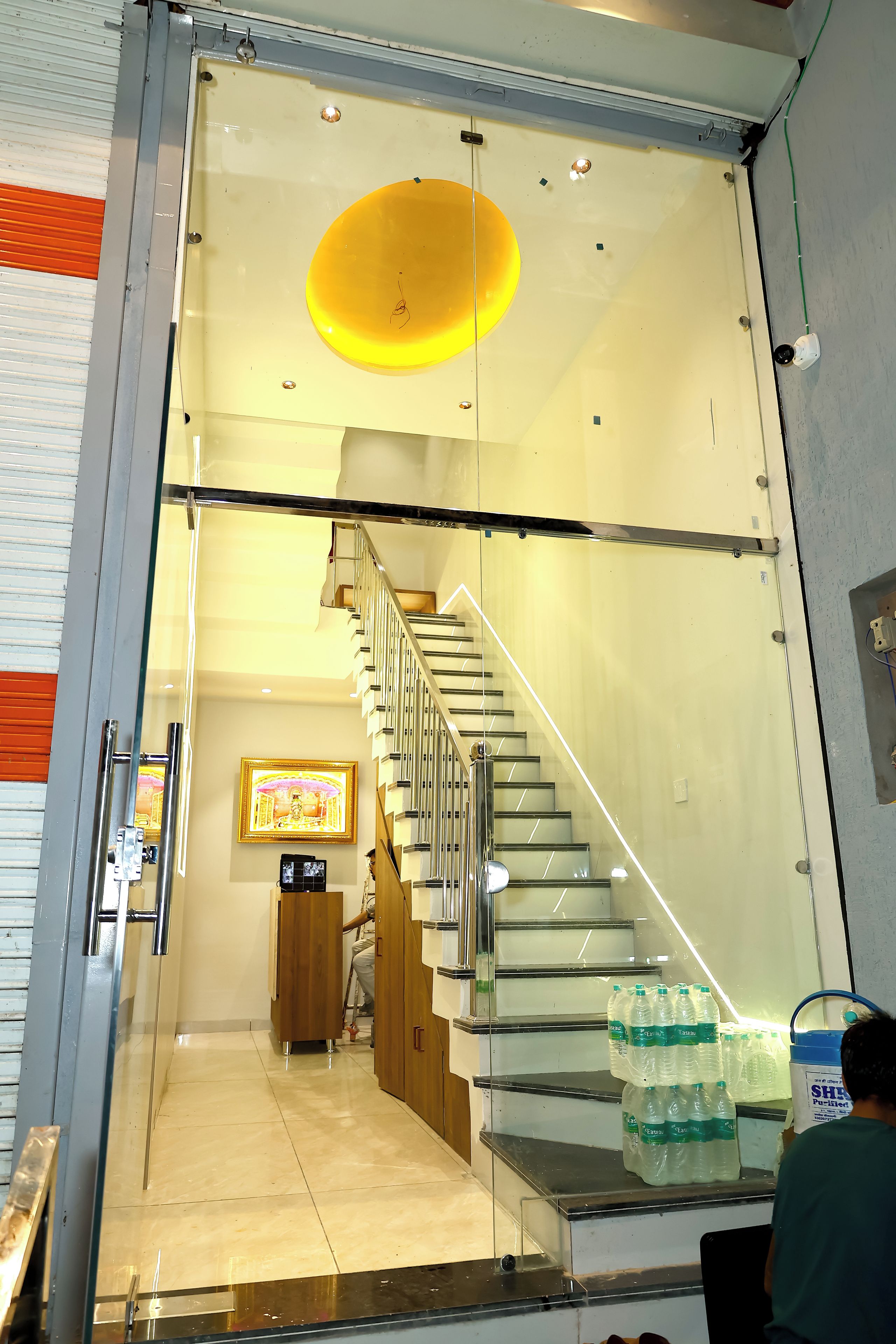 A modern interior with a glass staircase leading to a lower level, a framed picture on the wall, and bottled water on the floor.