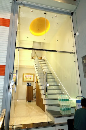 A modern interior with a glass staircase leading to a lower level, a framed picture on the wall, and bottled water on the floor. - Hotel Ishaan Palace (Ujjain)