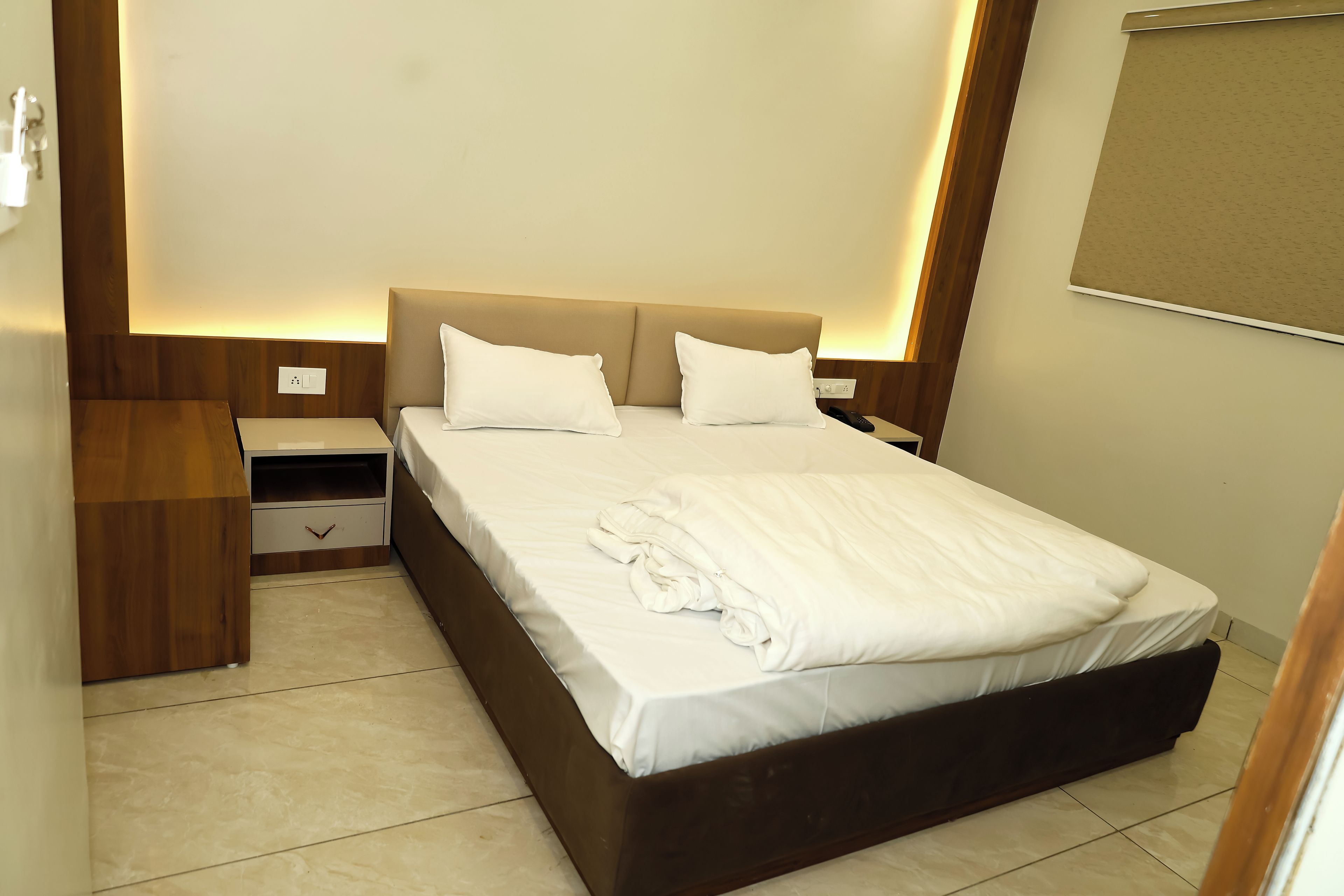 A neatly made bed with white linens and pillows, a wooden nightstand with a drawer, and a wall-mounted light fixture.