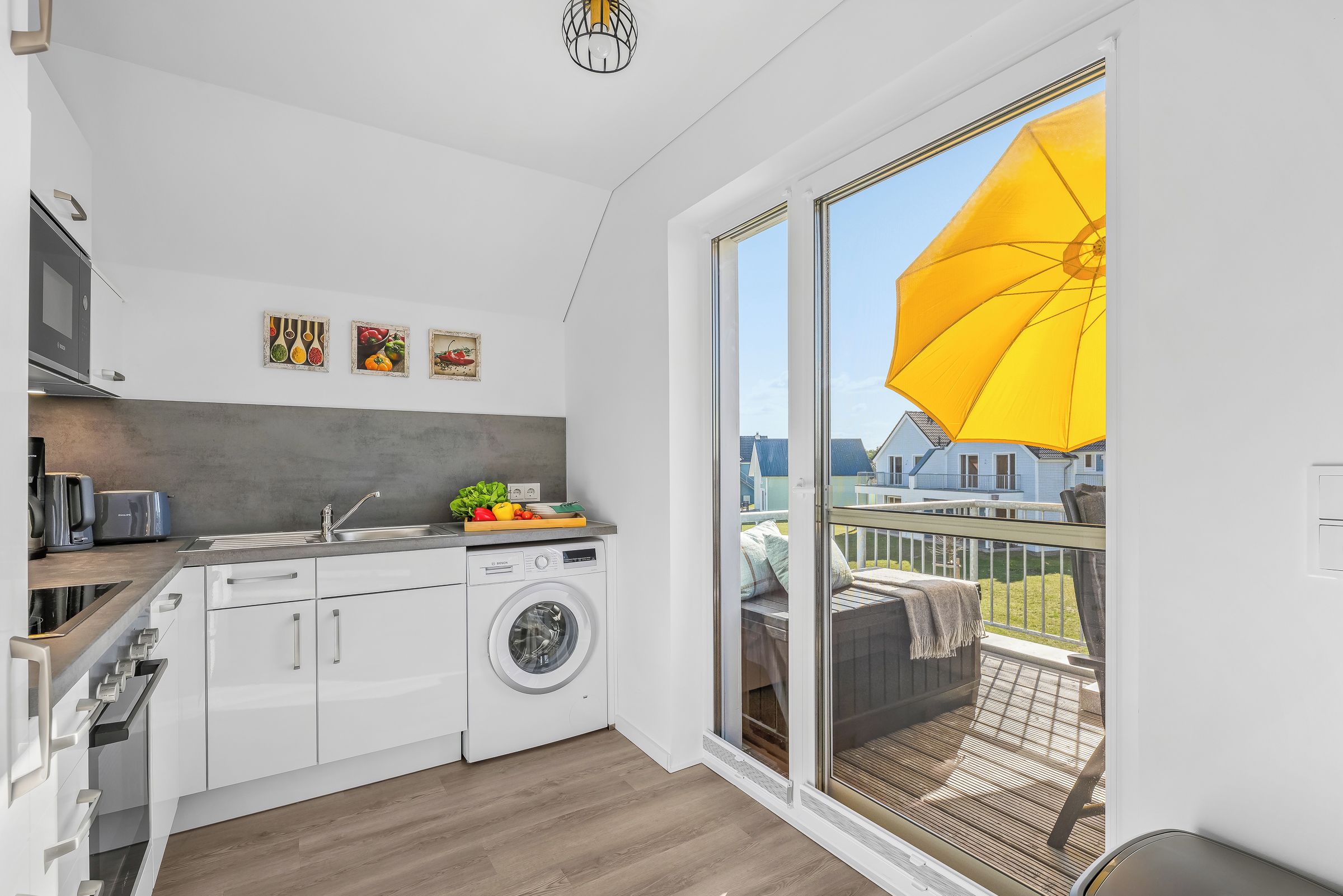A compact kitchen with a washing machine, a microwave, and a sink. There's a balcony with a yellow umbrella.