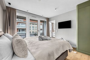 A modern bedroom with a large bed, a flat-screen TV mounted on the wall, and a balcony with a view of a building. - Westside Penthouse with a sauna, fireplace, balcony, and underground parking space (Sylt)