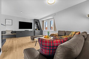 A modern living room with a flat-screen TV, grey sofas, a coffee table with fruits, and a window with curtains. - Eastside Penthouse with a sauna, fireplace, balcony, and underground parking space (Sylt)
