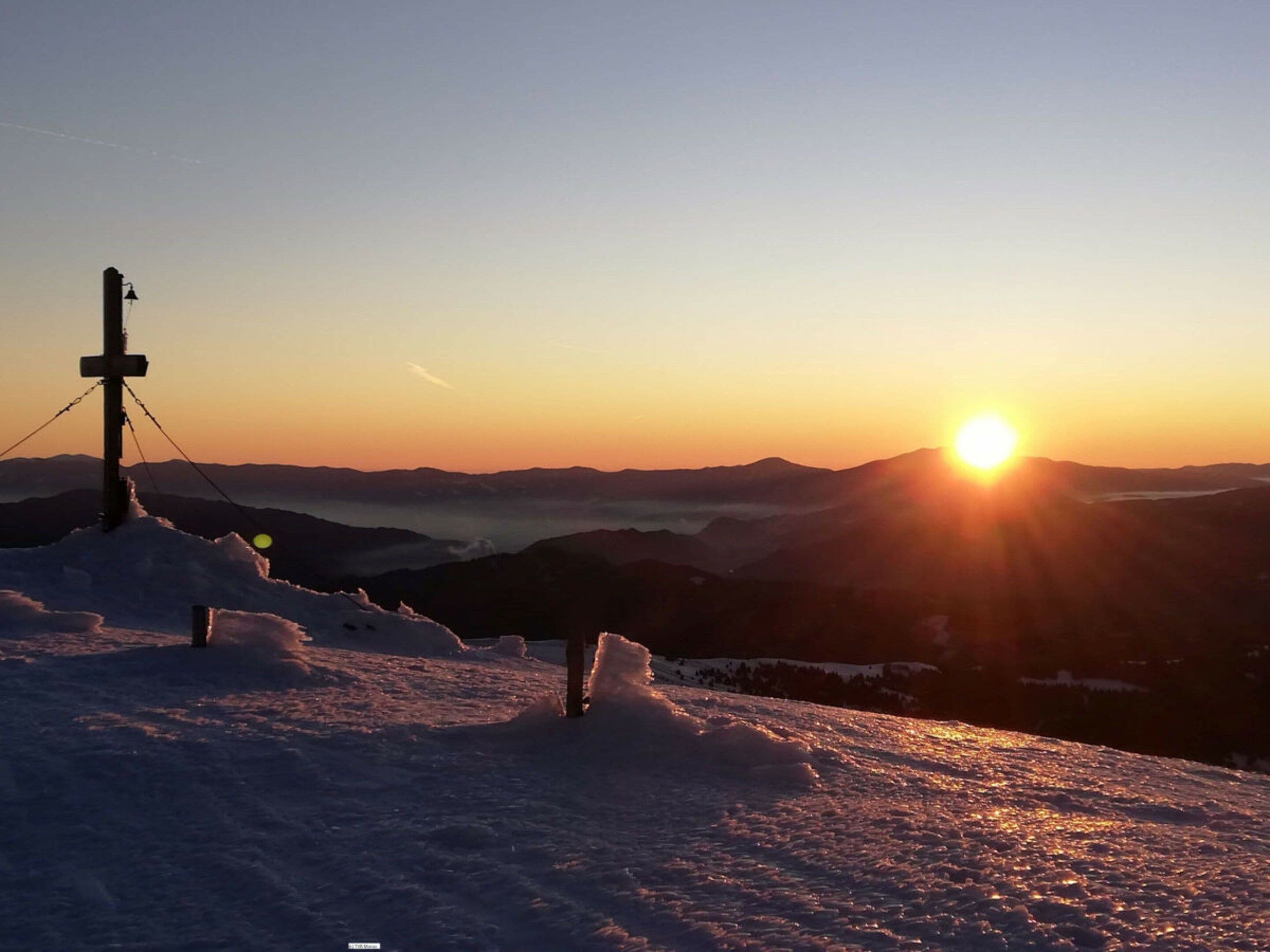 Un paysage enneigé avec une croix et un poteau téléphonique, des montagnes en arrière-plan, et le soleil se couchant ou se levant.