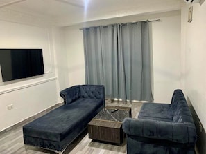 A living room with a sectional sofa, a coffee table, and a flat-screen TV mounted on the wall. - Contemporary City Flat with Comfortable Bedroom & Spacious Living Area (Accra)