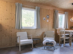 Interior de cabana de madeira com duas cadeiras, uma mesa pequena e uma janela com cortinas.