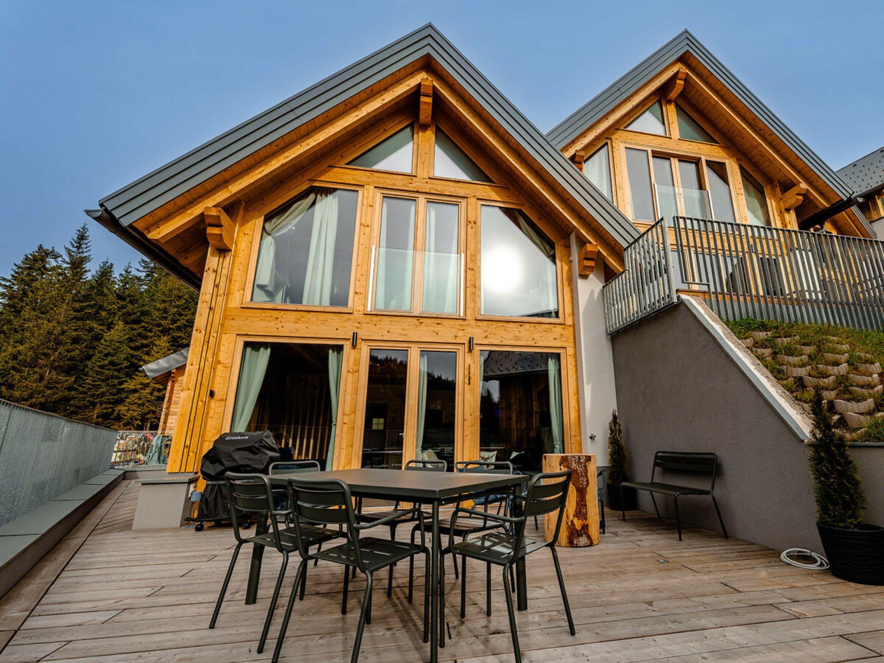 A wooden house with a balcony, a patio with a table and chairs, and a stone wall.