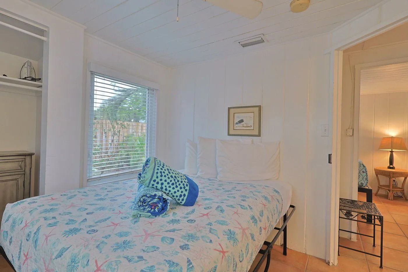A bedroom with a bed, a window with blinds, a bedside table with a lamp, and a door leading to another room.