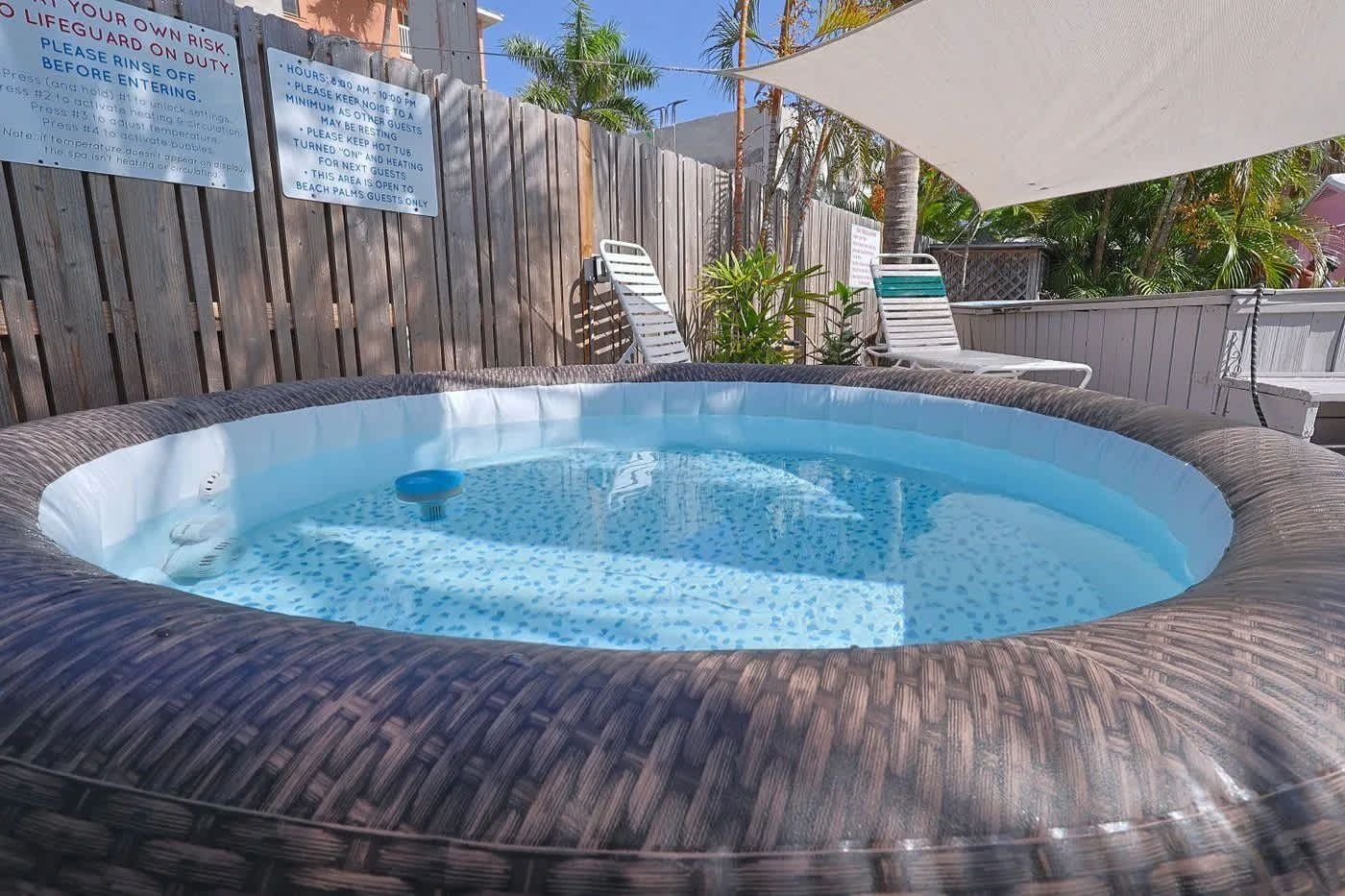 A hot tub surrounded by a wooden fence, with lounge chairs and an umbrella in the background.