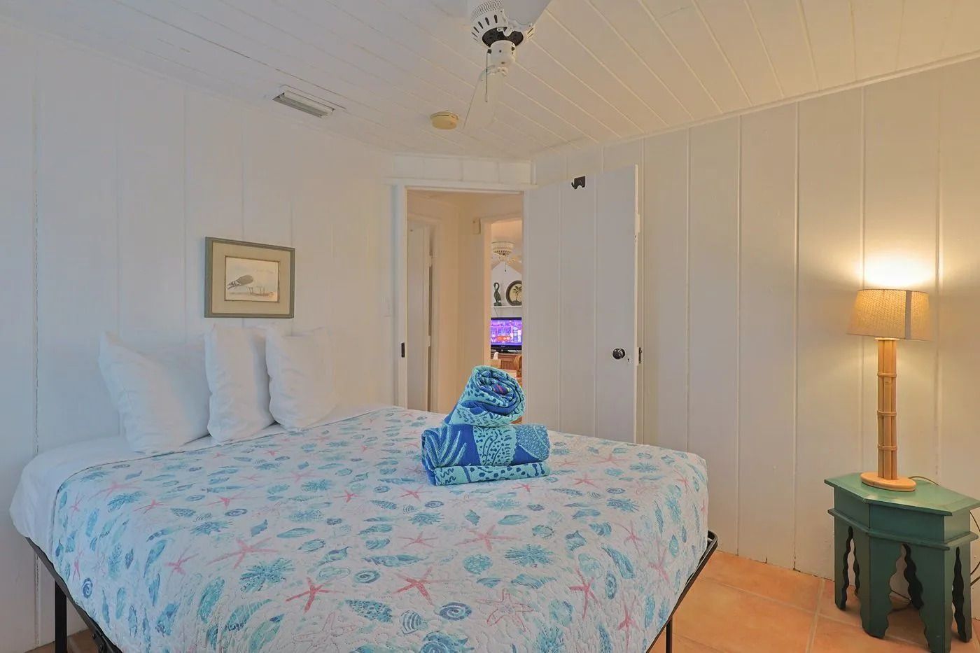A bedroom with a bed, a framed picture on the wall, a door leading to another room, and a bedside lamp.