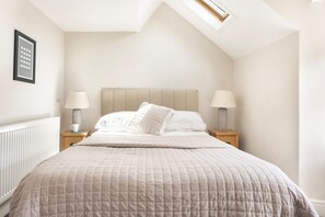 Un dormitorio con techo inclinado, una cama con colcha acolchada color beige, dos mesitas de noche con lámparas y un cuadro en la pared.