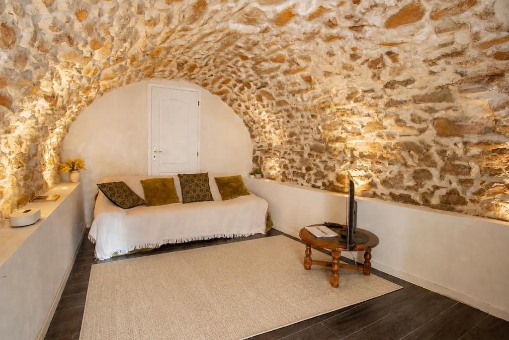 Una habitación acogedora con pared de piedra, un sofá blanco con cojines, una pequeña mesa de madera y una puerta blanca.