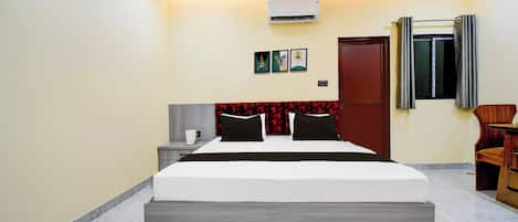 A hotel room with a bed, bedside table, wall-mounted air conditioning unit, and a door with a window.