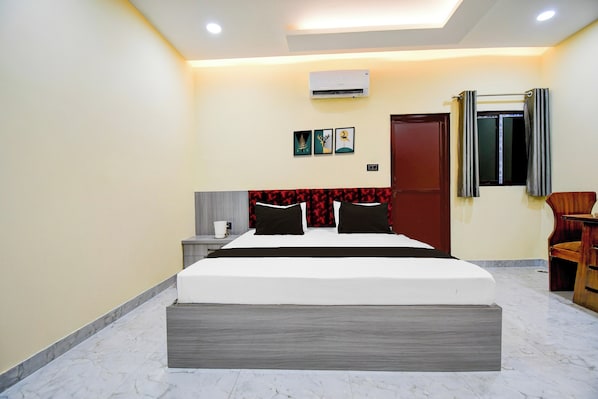 A hotel room with a bed, bedside table, wall-mounted air conditioning unit, and a door with a window.
