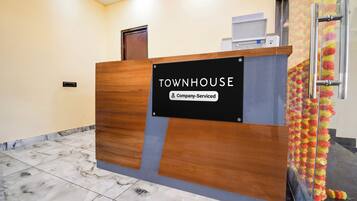 Un mostrador de recepción con la etiqueta 'TOWNHOUSE' y un cartel que indica 'Servicio de empresa', una puerta, un escritorio pequeño y un teléfono instalado en la pared.