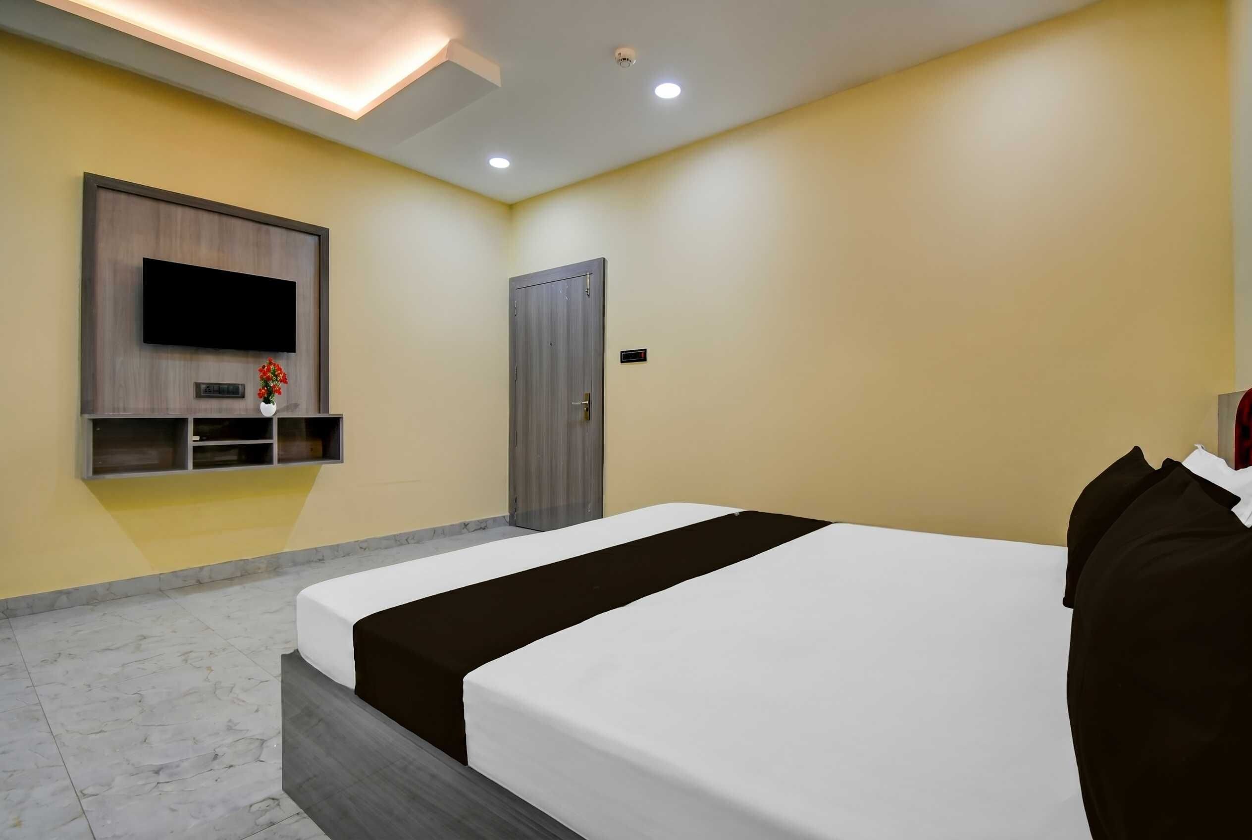 Habitación de hotel con una cama, televisión montada en la pared, una puerta y un pequeño estante con un florero.