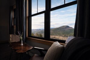A room with a large window, a wooden table with a vase, and a cushioned bench. - Luxury Mountain Escape • 6BR • Hot Tub & Views • 4mi to Sunday River (Bethel)