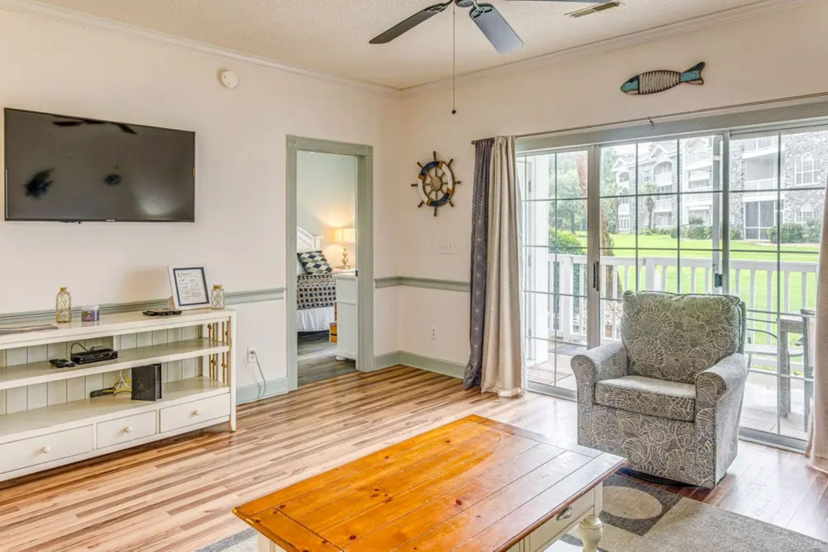 Un salon avec une table basse en bois, un téléviseur fixé au mur, un fauteuil confortable et une grande fenêtre donnant sur une maison et une pelouse.