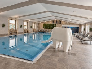 Una amplia piscina cubierta con un fondo de pared de piedra, sillas de descanso y un tobogán acuático con forma de elefante.