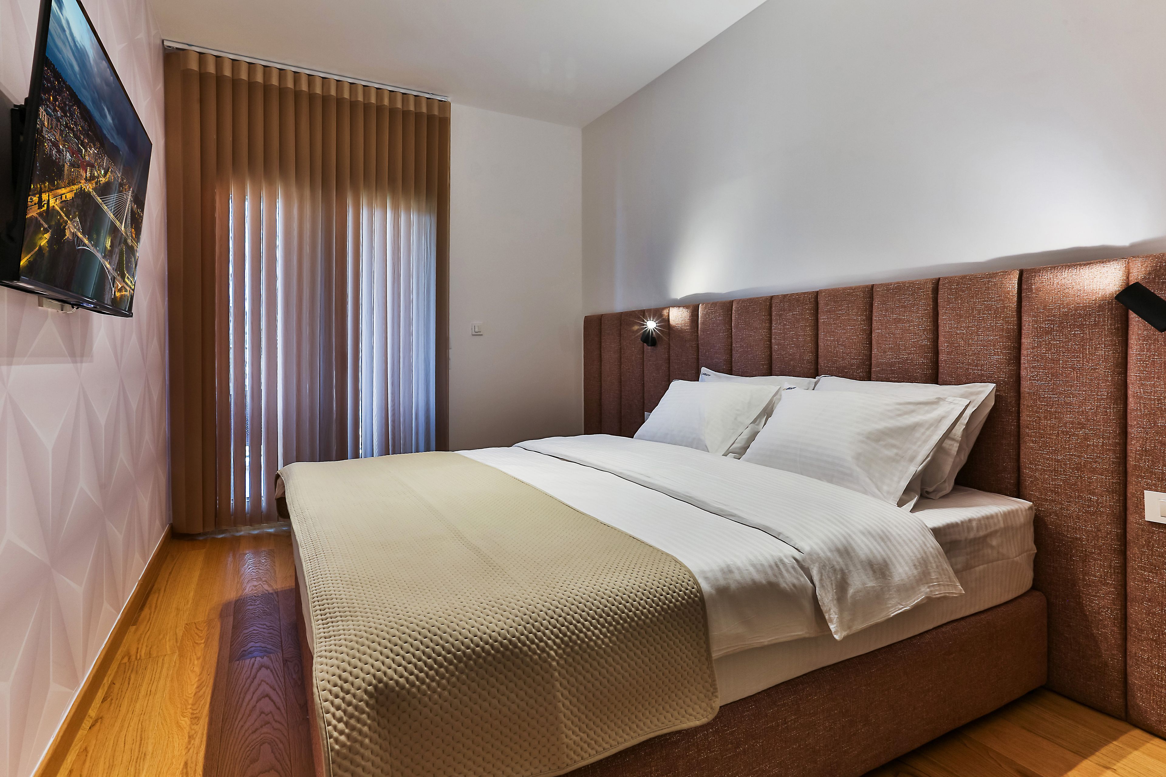 Un dormitorio moderno con una cama grande, cabecera con iluminación integrada, televisor montado en la pared y ventana con persianas.