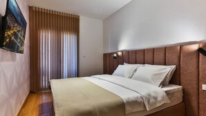 Een moderne slaapkamer met een groot bed, een hoofdbord met ingebouwde verlichting, een wandtelevisie en een raam met zonwering.
