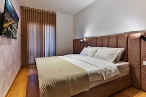 Un dormitorio moderno con una cama grande, cabecera con iluminación integrada, televisor montado en la pared y ventana con persianas.