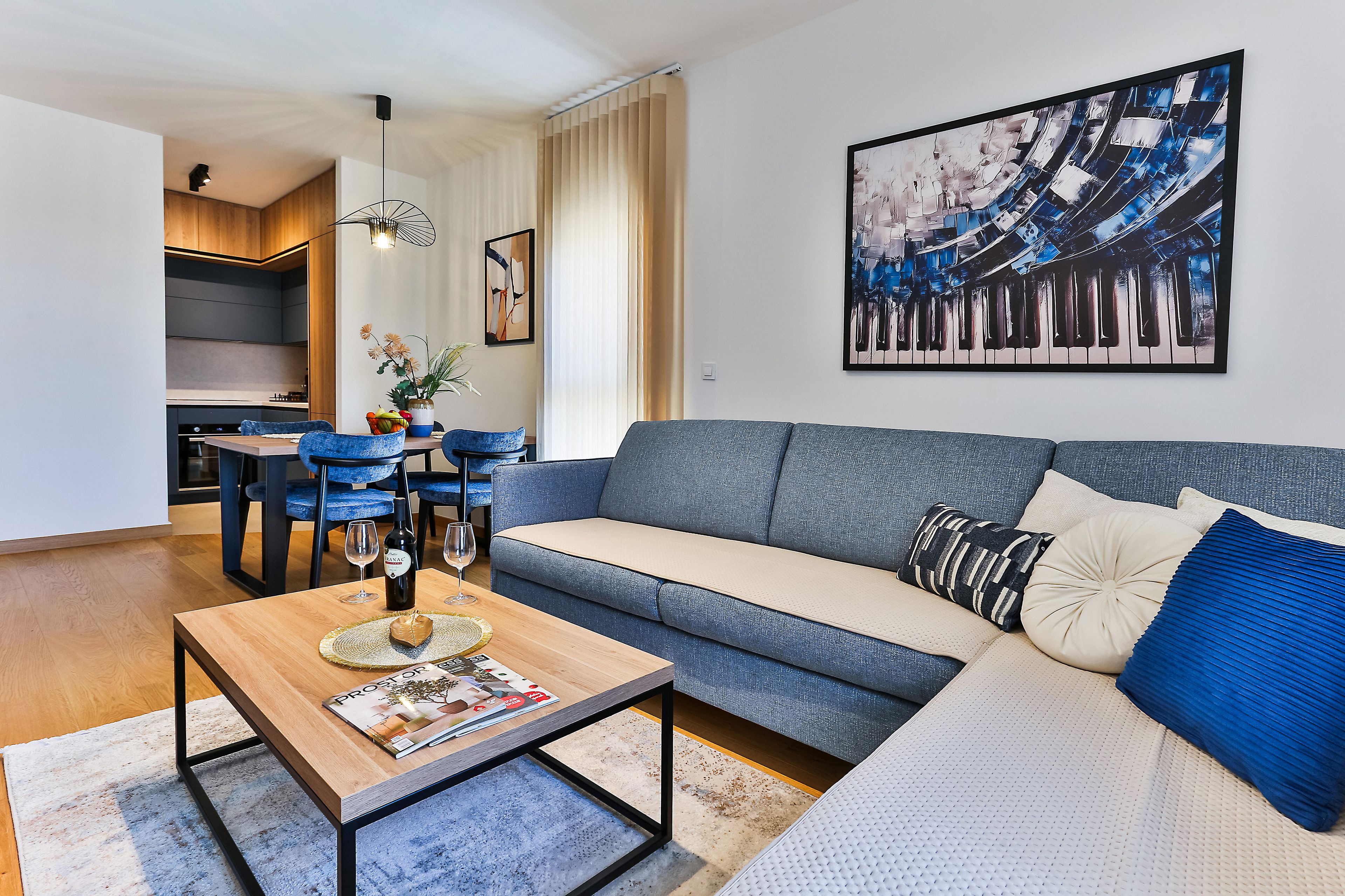 A modern living room with a grey sofa, a wooden coffee table with a magazine, and a dining area with blue chairs and a vase of flowers.