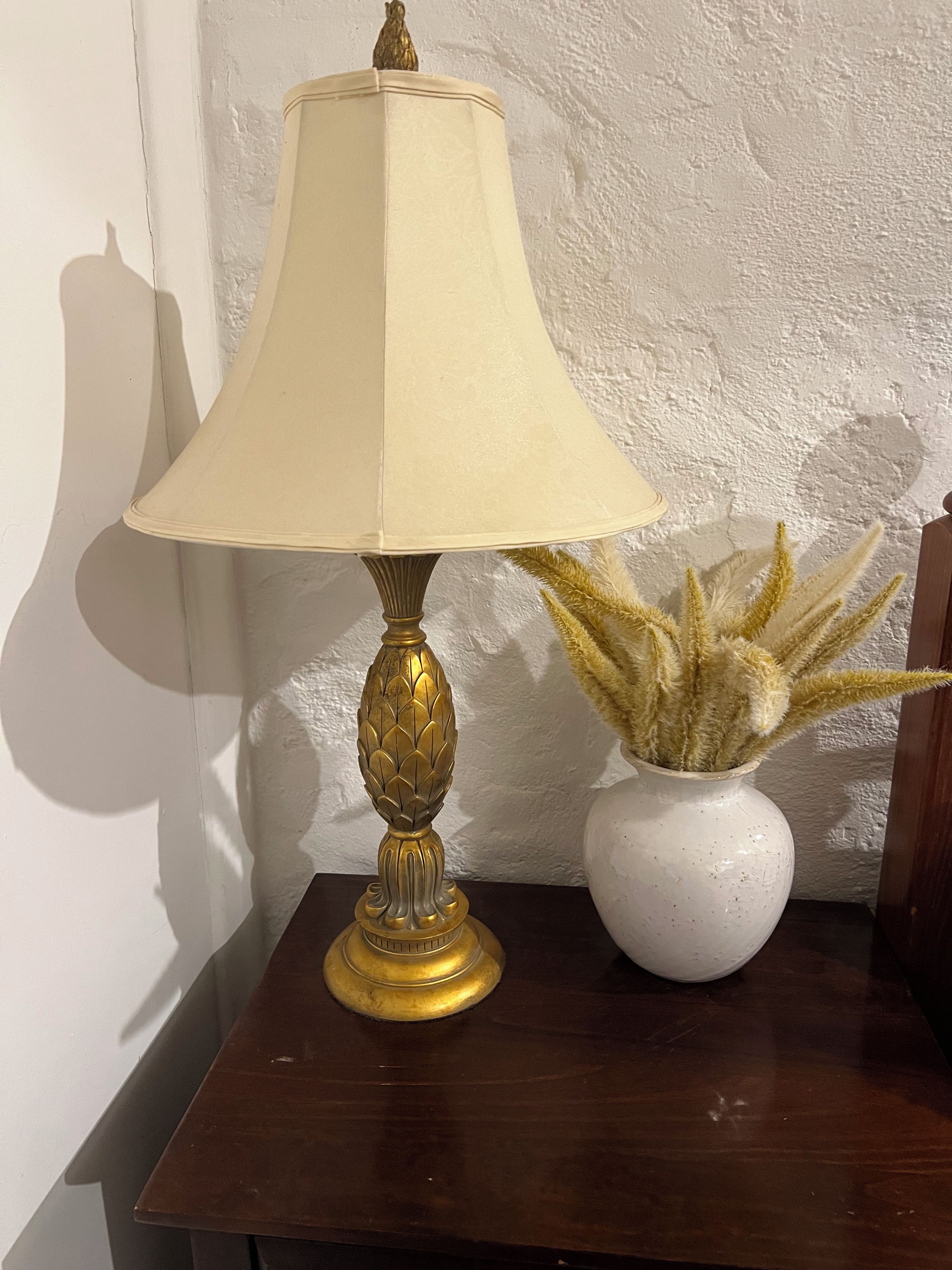 Une lampe avec un abat-jour beige et un pied décoratif, posée sur une table en bois à côté d’un vase blanc contenant des plantes séchées.