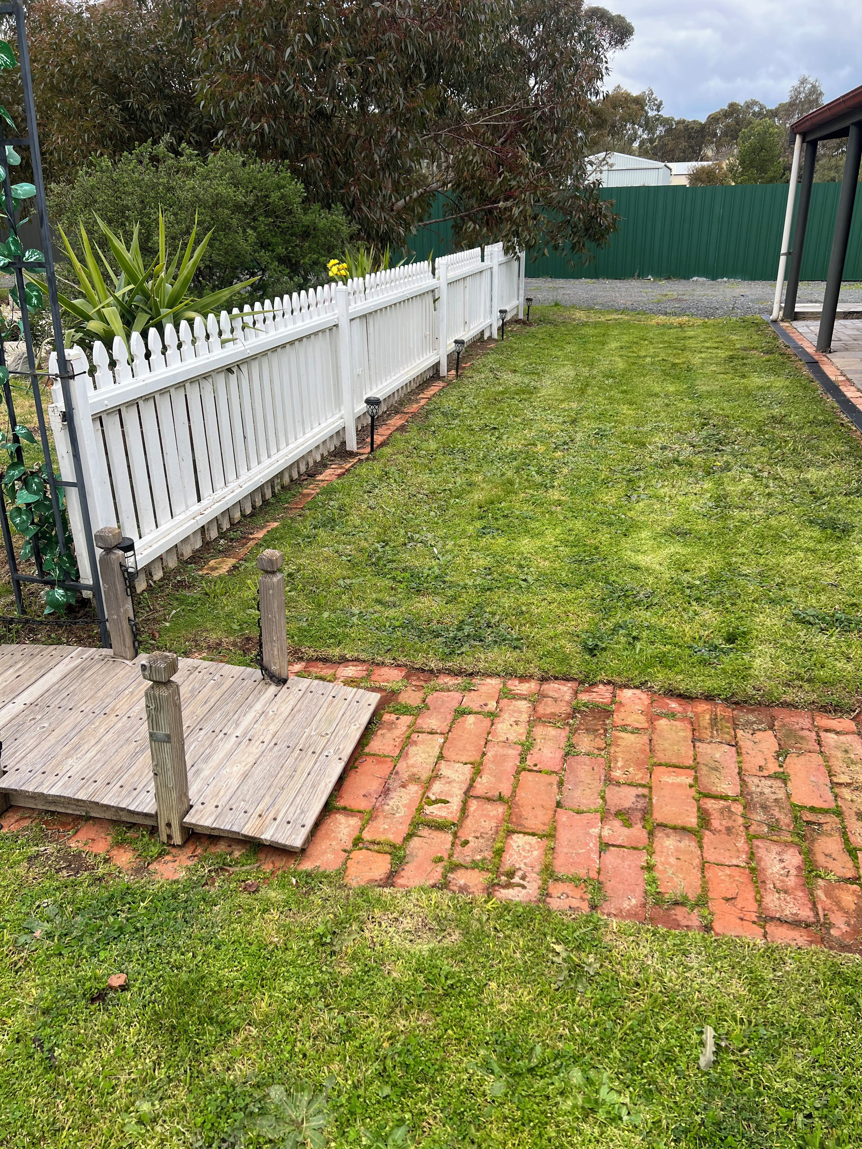 A paved pathway leading to a wooden deck with a white picket fence and a green fence in the background.