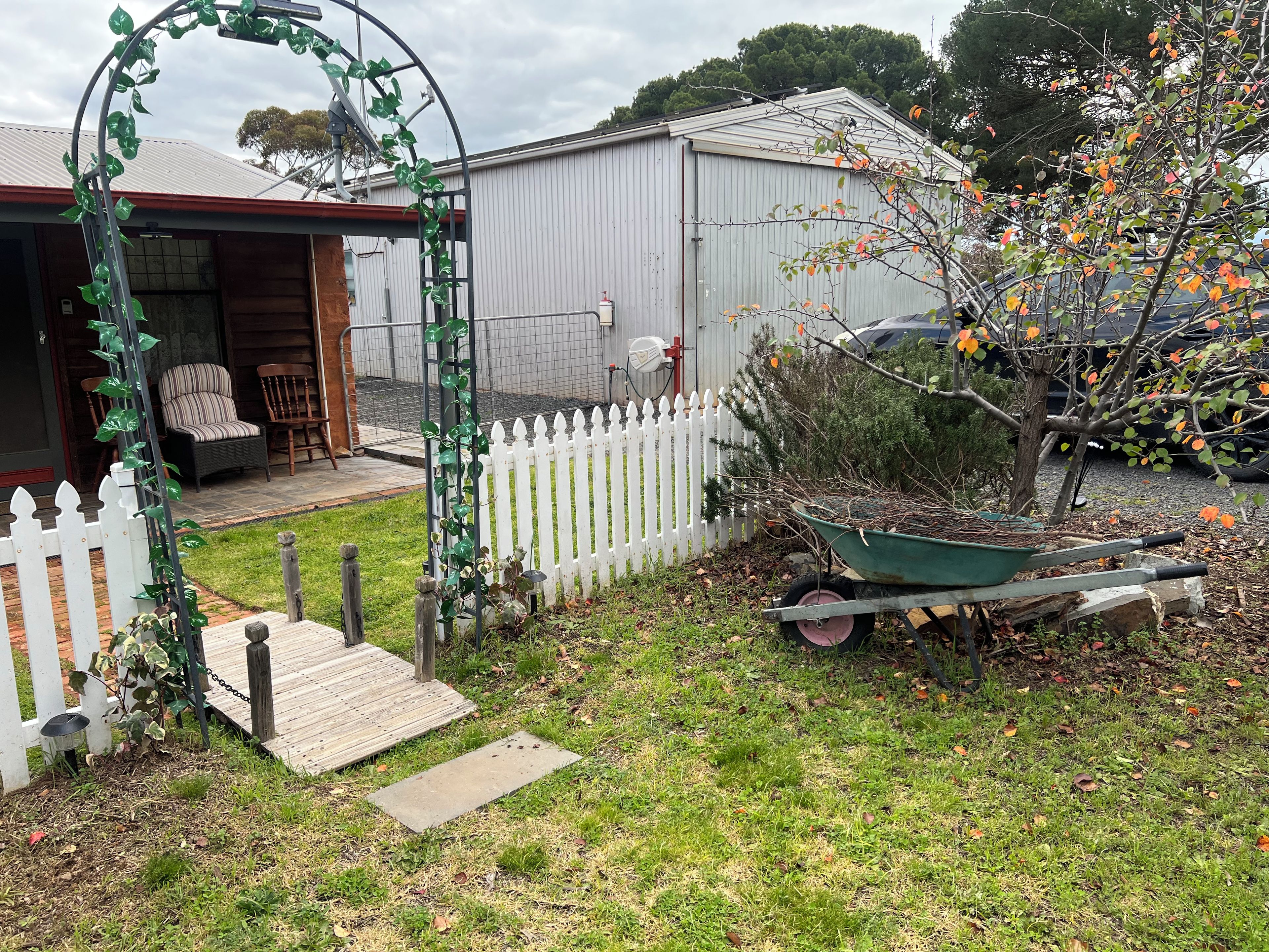A residential backyard with a wooden deck, a white picket fence, a wheelbarrow, and a garden area.