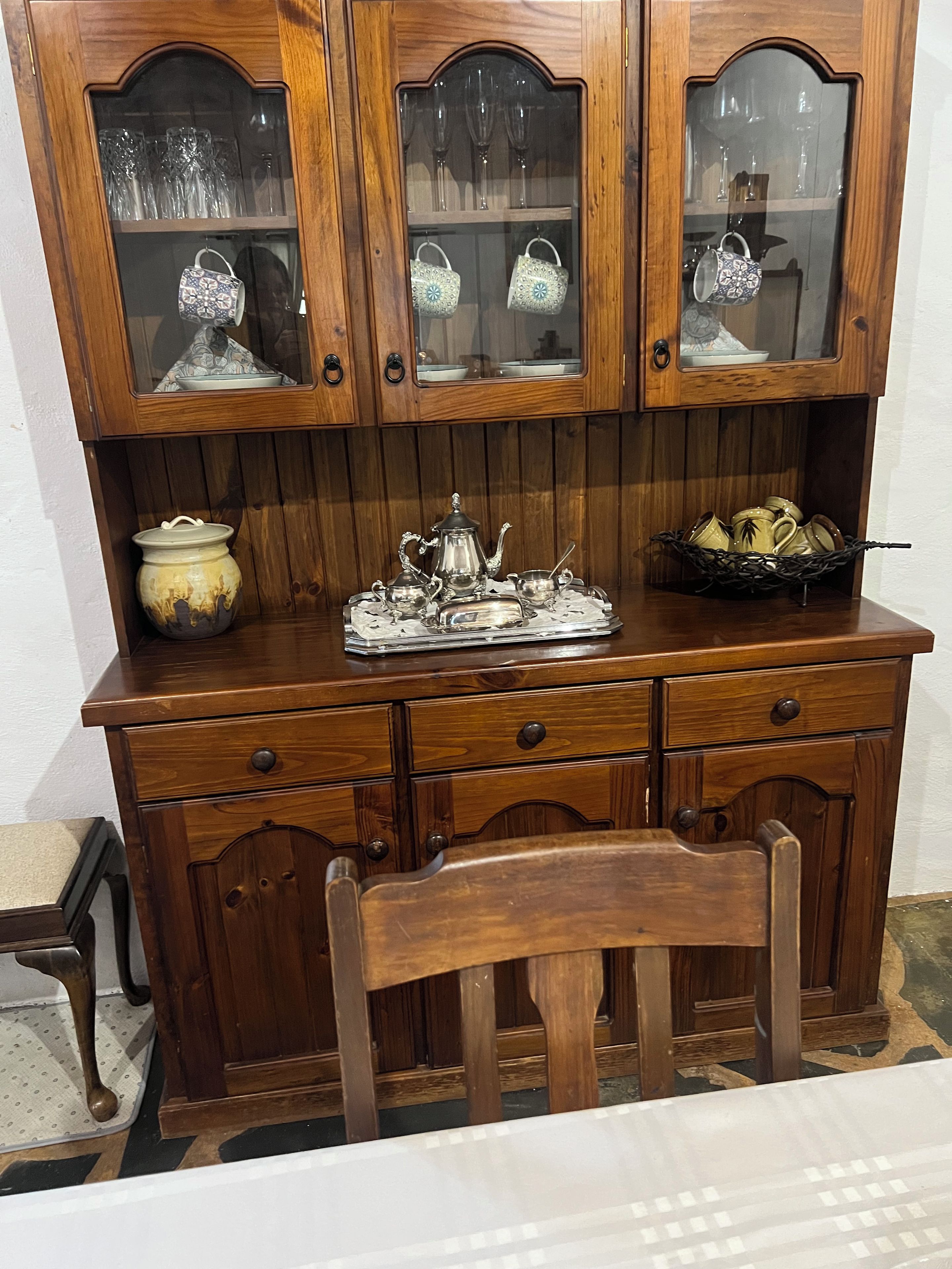 A wooden cabinet with glass-fronted compartments containing glassware and a tray with silverware.