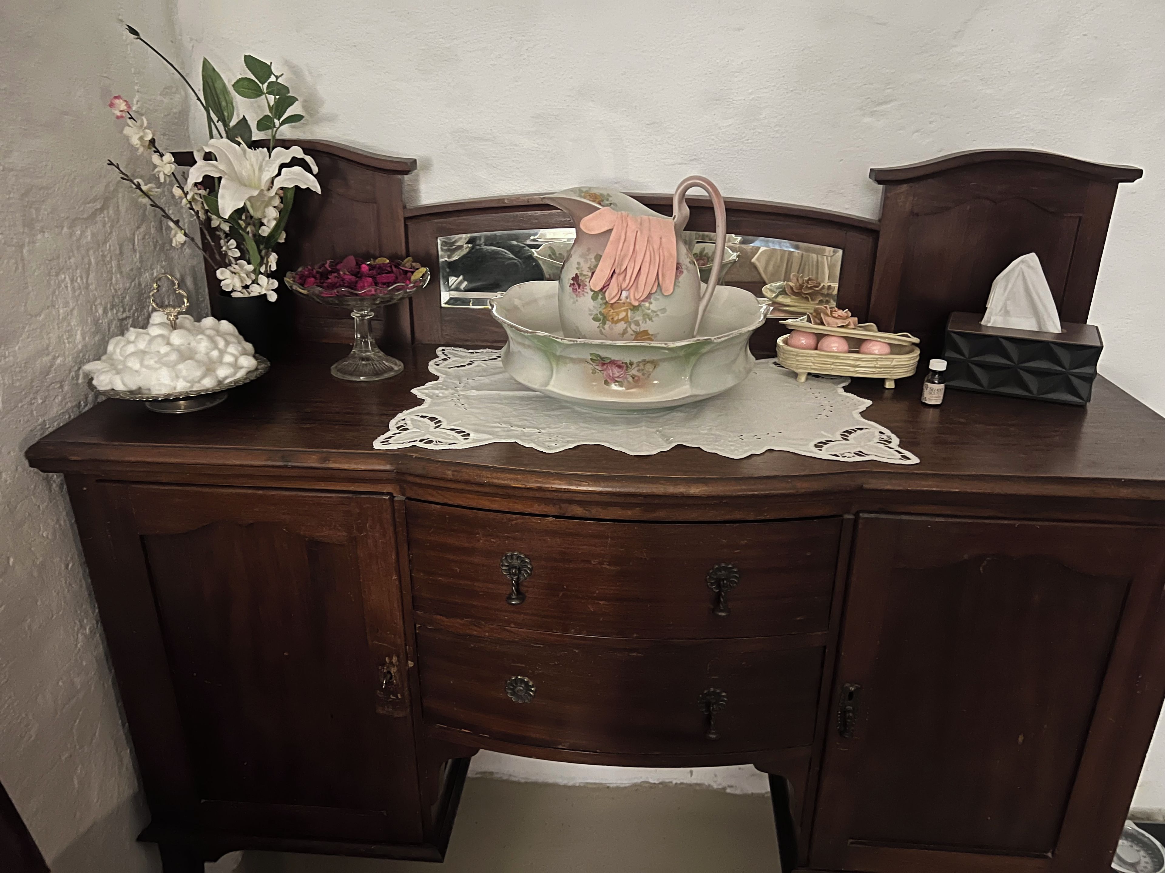 Ein hölzerner Nachttisch mit einer Spitzenerdecke, eine Schale mit weißen, kugelförmigen Objekten, eine Vase mit Blumen und eine Taschentuchbox.