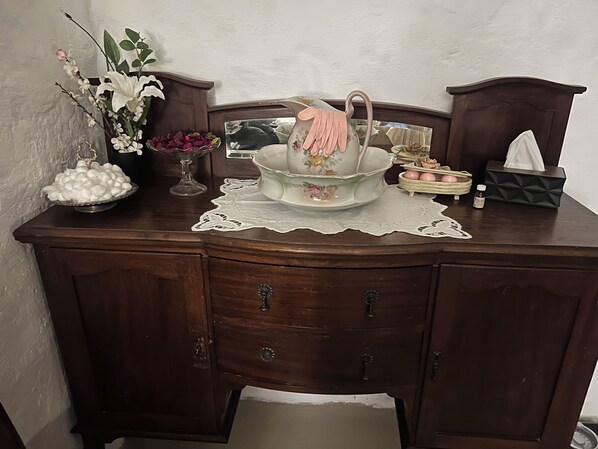 A wooden dresser with a lace doily, a bowl of white spherical objects, a vase with flowers, and a tissue box.