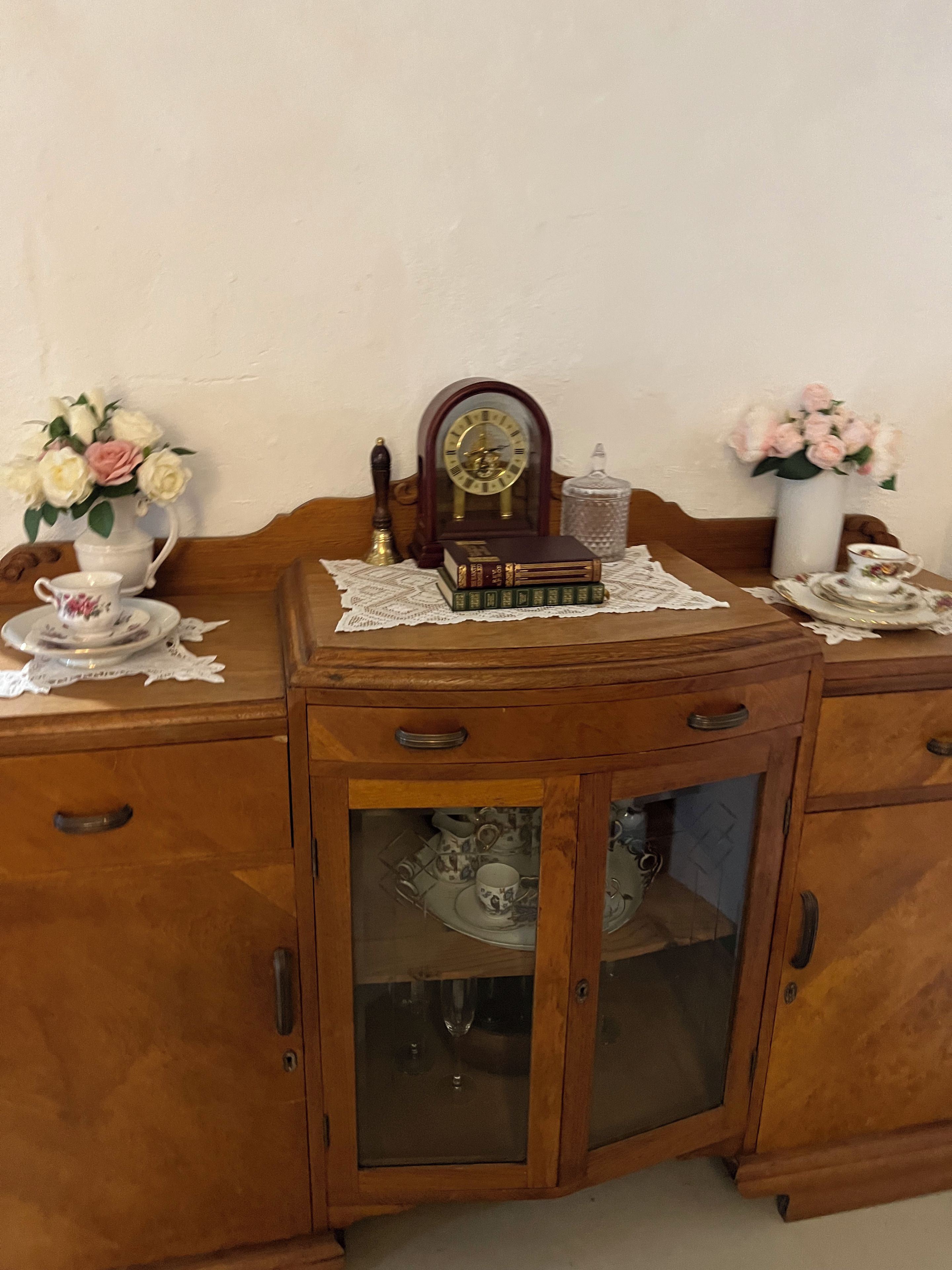 A wooden cabinet with a glass-fronted cabinet drawer, a clock, books, a teapot, and a vase with flowers.