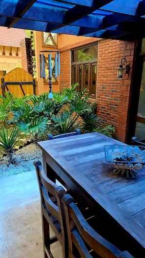 Eine Terrasse im Freien mit einem Holztisch und Stühlen, umgeben von Grünpflanzen und einer Ziegelwand.