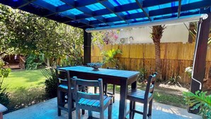 Een overdekt terras met een tafel en stoelen, omgeven door een houten omheining en groen.
