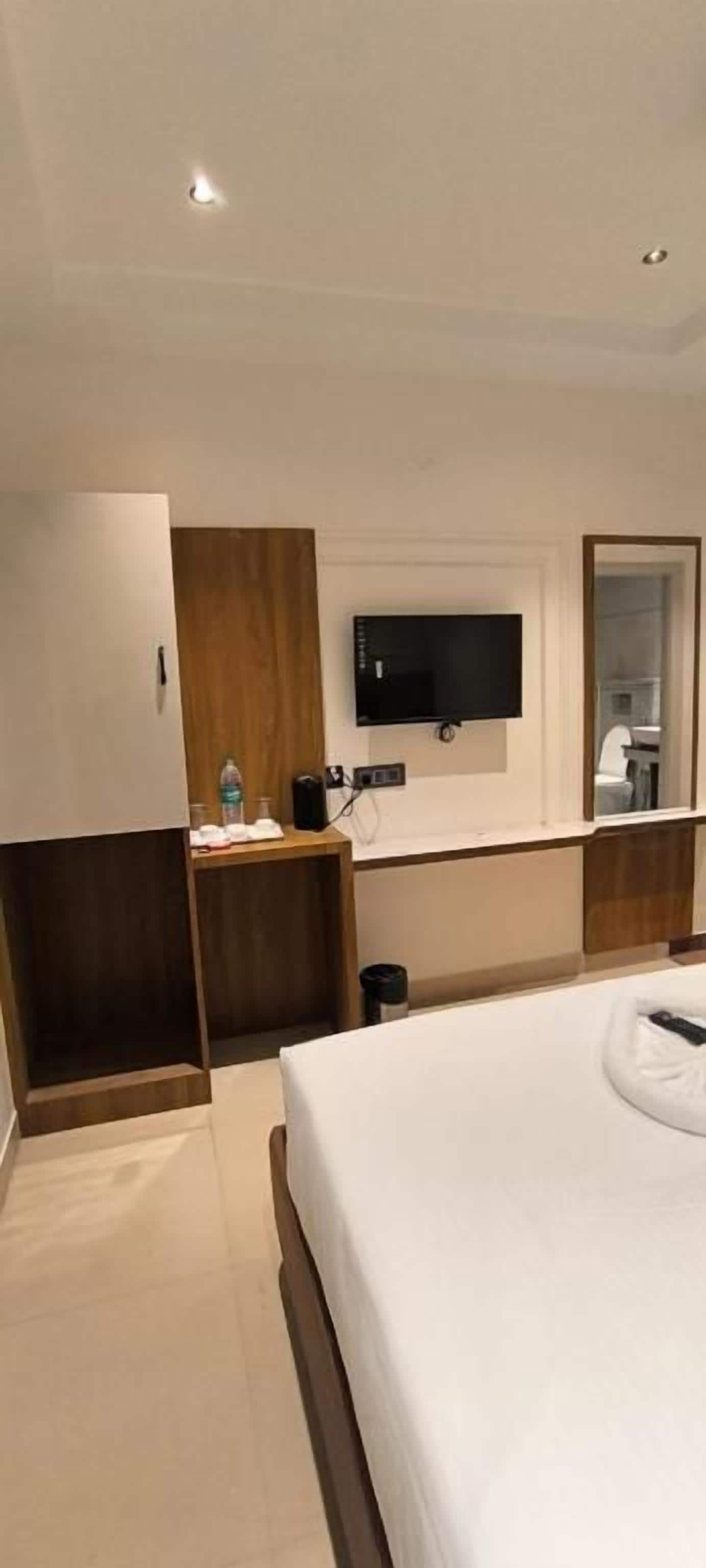 Habitació d'hotel amb un llit, un televisor fixat a la paret, un armari de fusta i una taula petita amb una ampolla i una tassa.