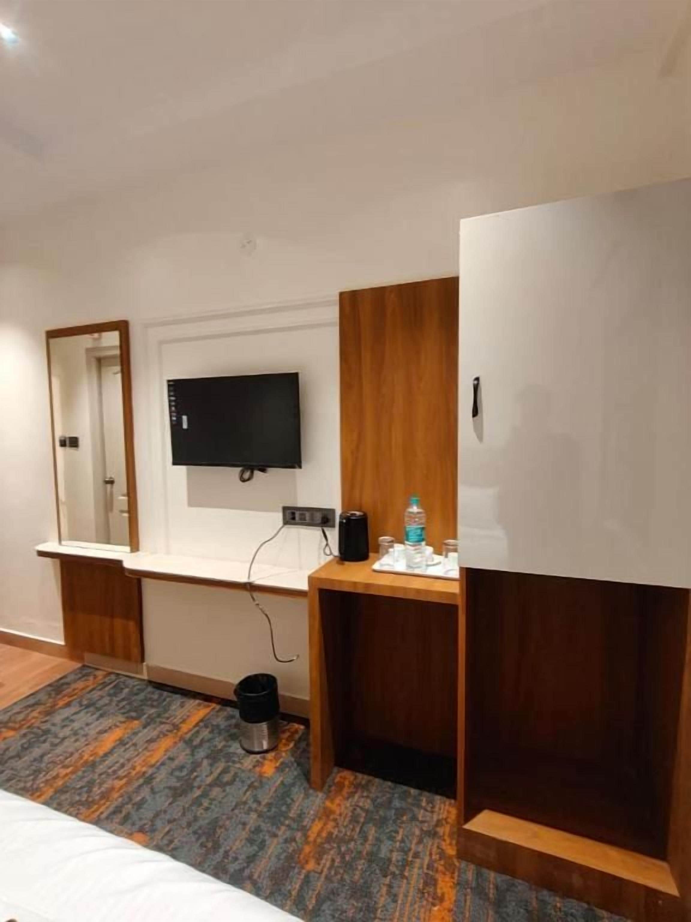 A hotel room with a bed, a desk with a flat-screen TV, a mirror, and a small trash bin.