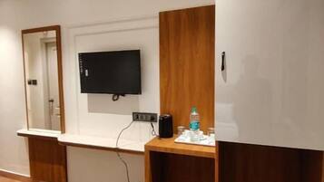 A hotel room with a bed, a desk with a flat-screen TV, a mirror, and a small trash bin.