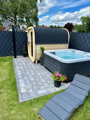 A hot tub with a wooden exterior, surrounded by a dark fence, a lounge chair, and potted plants.