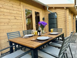 A wooden table set for two with chairs, a bottle of wine, a glass, and a vase with purple flowers.