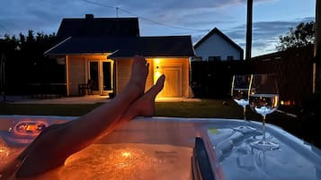 Iemand ontspant in een hottub met een glas wijn, met uitzicht op een achtertuin met een houten huis en een hek.