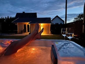 A person relaxing on a hot tub with a glass of wine, overlooking a backyard with a wooden house and a fence.